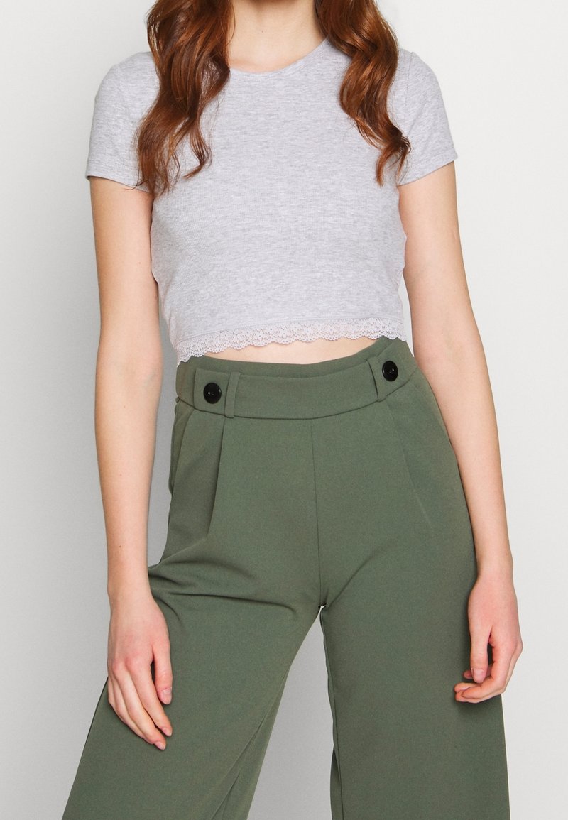 Femme portant un top court gris clair à manches courtes avec bordure en dentelle et un pantalon taille haute vert olive avec des boutons noirs.