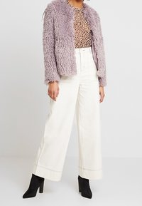 Väärennetut turkisjakku vaalean violetissa, täplikäs yläosa beige- ja ruskeakuosisena, leveät valkoiset housut ja mustat korolliset nilkkurit.