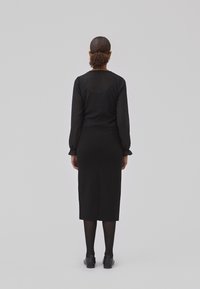 Blouse noire à manches longues avec un motif texturé, associée à une jupe crayon noire. Le modèle porte des collants et des chaussures noires, vu de dos.