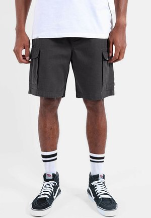 Homme portant un short cargo noir, des chaussettes blanches avec des rayures noires et des baskets montantes noires avec des lacets blancs, debout devant un fond uni.