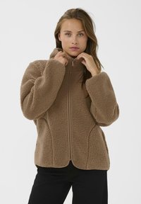 Barna fleece dzseki magas gallérral, cipzáras záródással és strukturált anyaggal; laza szabású, kontrasztos szegéllyel az illesztéseken.