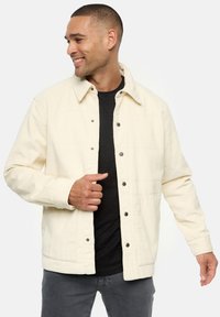 Cremefarbene Cordjacke mit Kragen, Druckknöpfen, zwei seitlichen Taschen und strukturiertem Stoff. Getragen über einem schwarzen Hemd und grauen Hosen.