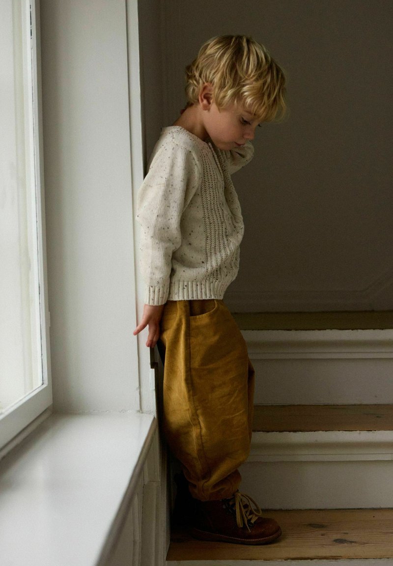 Enfant aux cheveux blonds portant un pull beige et un pantalon moutarde, debout près d'une fenêtre et d'un escalier, regardant vers le bas avec une main derrière le dos.
