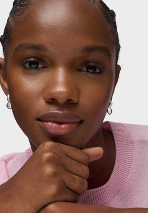 Zbliżenie osoby o ciemnej karnacji, opierającej podbródek na pięści, noszącej srebrne kolczyki i jasnoróżowy sweter na jednolitym tle.