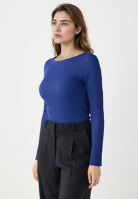 Niebieska, prążkowana bluzka z długim rękawem, okrągłym dekoltem i dopasowanym krojem. Połączona z ciemnymi spodniami z wysokim stanem. Gładkie tło.
