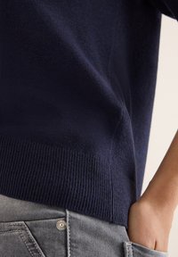 Marineblauer Strickpullover mit geripptem Saum, getragen über grauen Jeans. Weiche Textur sichtbar, Hand in der Tasche ruhend, was den Lagenstil zeigt.