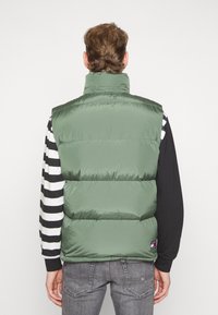 Zelená prošívaná vesta s vysokým límcem, prošitým vzorem a viditelnými švy. Nositelná přes černobíle pruhované tričko s dlouhým rukávem.