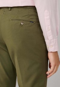 Pantalones verde oliva con una tela texturizada, que incluyen un bolsillo trasero con botón y una etiqueta de la marca; combinados con una camisa de manga larga de color rosa claro.
