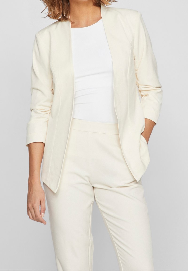 Femme portant un blazer crème sur mesure avec des manches trois-quarts, un pantalon crème assorti et un haut blanc ajusté, debout avec une main dans la poche.
