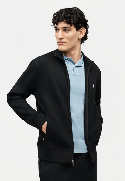 Mladý muž s krátkými kudrnatými vlasy, oblečený v světle modrém polotričku a černé bundě na zip, s rukama v kapse, dívající se stranou.