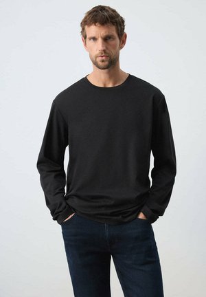 Hombre con cabello corto y castaño, vestido con una camiseta negra de manga larga y vaqueros azul oscuro, de pie sobre un fondo claro y liso.