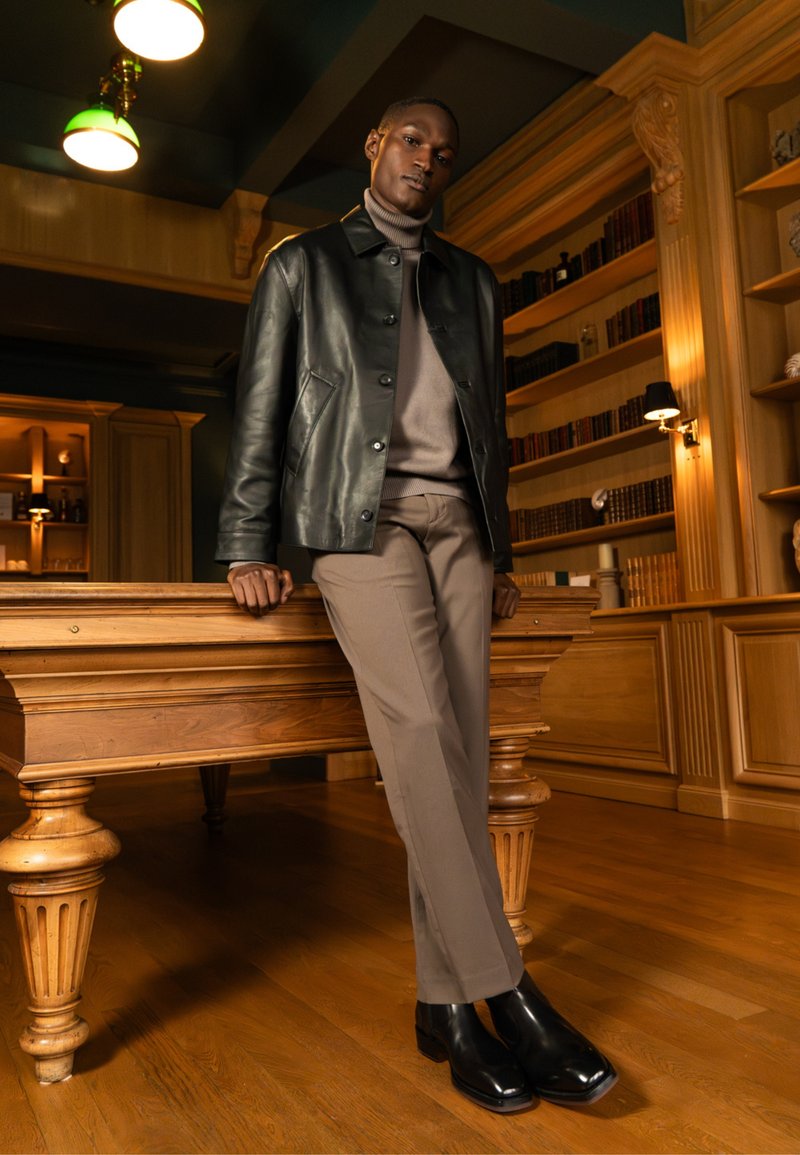 Homme appuyé sur une table de billard en bois dans une pièce avec des étagères remplies de livres, portant une veste en cuir noire, un col roulé marron, un pantalon beige et des bottes noires.