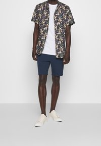 Chemise à manches courtes à motifs floraux dans des couleurs sombres avec des accents roses et verts, associée à un short marine et des baskets beige. Ajustement simple et décontracté.