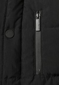 Czarny materiał kurtki puchowej z pikowaną fakturą. Zawiera kieszeń na zamek błyskawiczny z czarnym uchwytem oraz okrągły guzik z logo marki.