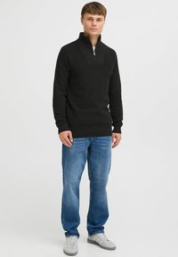 Pulover negru cu guler cu fermoar, din tricot texturat, cu mâneci lungi și tiv cu striații, asortat cu blugi albaștri și adidași gri pe un fundal neutru.