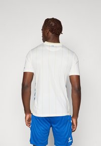 Hvid atletisk trøje med beige lodrette striber, korte ærmer, rund halsudskæring og blå detaljer; parret med blå shorts.