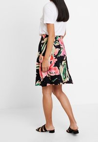 Jupe florale en tissu noir ornée de fleurs roses et vertes, design en A, mi-longue, associée à un t-shirt blanc et des sandales noires.