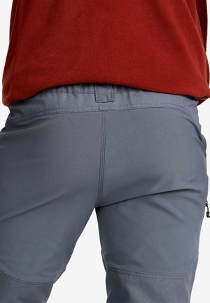 Närbild av en person som bär grå byxor och en röd fleecjacka, som visar sömnads- och midjedetaljer.