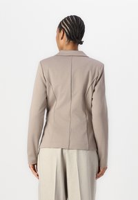 Přiléhavý blazer s dlouhým rukávem v barvě světle taupe, vyrobený z hladké tkaniny. Má klopový límec a strukturované švy na zadní části.