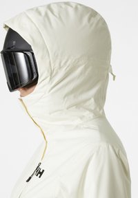 Crème-kleurige skijas met een hoge kraag, capuchon en ritssluiting. Kenmerkt zich door een gladde textuur en een bedrukt logo op de borst.