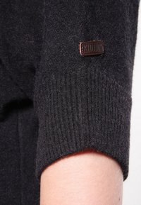 Tmavě šedý pletený svetr s žebrovanou texturou na manžetách, se značkovým štítkem z hnědého kovu. Tkanina vypadá měkká a mírně chlupatá.