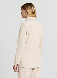 Blazer beige à côtes avec un col ajusté, des manches longues et des accents de poignet. Texture lisse et design minimaliste sans fermetures visibles.