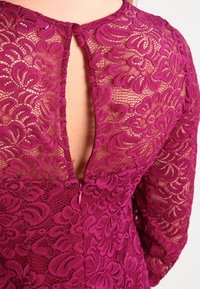 Magenta spetsklänning med blommönster, långa ärmar samt en nyckelhålsspringa bak med knappstängning och blixtlåsdetalj.