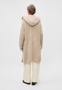 Manteau à capuche long en tricot beige avec une finition texturée, coupe décontractée et fentes sur les côtés, associé à un pantalon large crème et des chaussures noires.