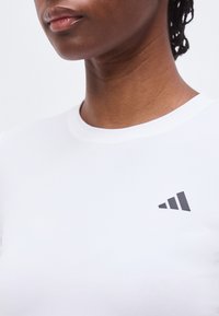 Gros plan d'une personne portant un T-shirt blanc avec un petit logo Adidas noir sur le haut de la poitrine.