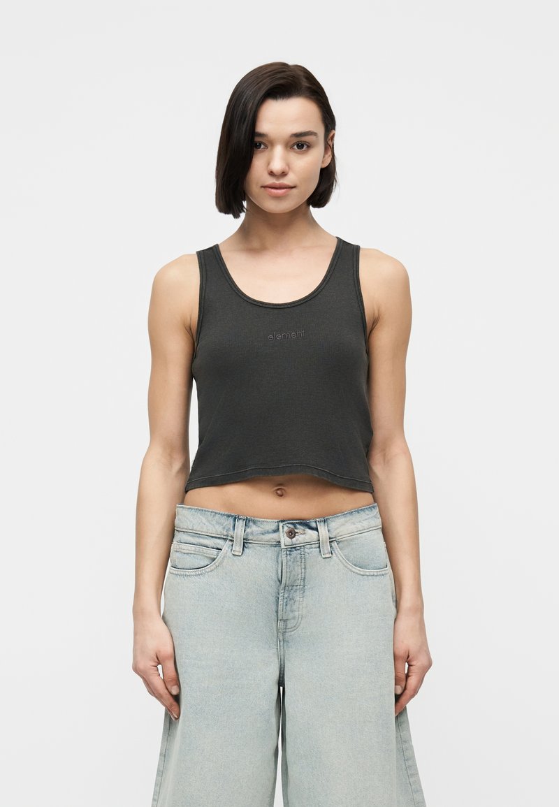 Jeune femme aux cheveux courts et foncés portant un débardeur noir court et un jean taille haute bleu clair à jambes larges, debout devant un fond blanc.