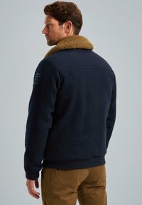 Námořnická fleece bunda s velkým hnědým beránkovým límcem, žebrovanými manžetami a lemem, a decentním detailem na ramenou. Nosená s khaki kalhotami.