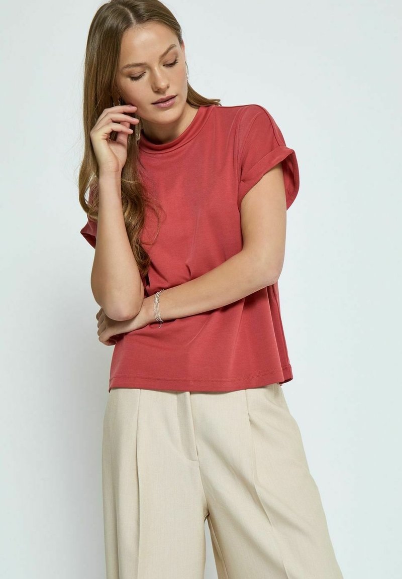 Rožinė trumpų rankovių palaidinė iš minkštos medžiagos su pririštomis rankovėmis, derinama su šviesiai smėlio spalvos aukšto liemens kelnėmis su raukšlėmis.