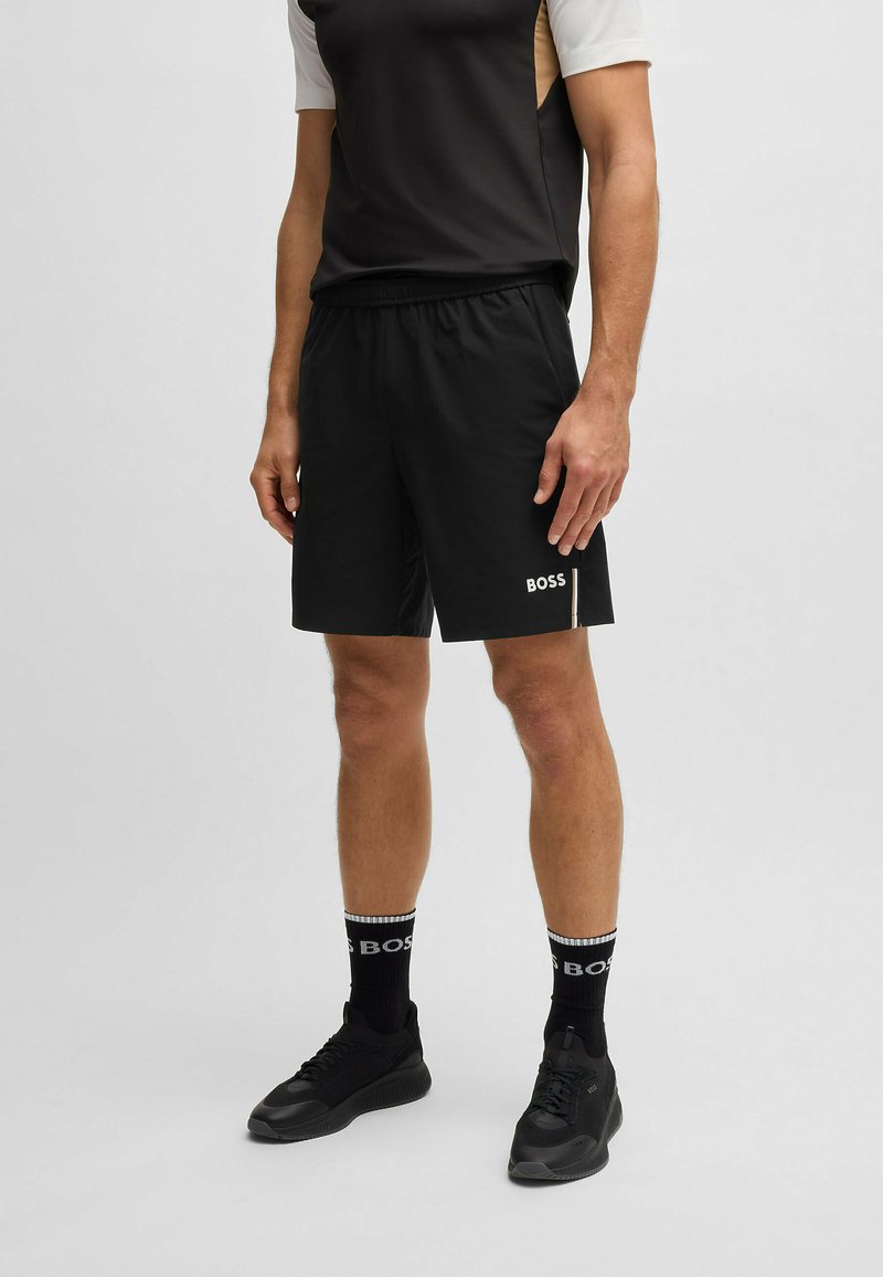 Melnas sportiskas šortus ar elastīgu jostasvietu, ar baltu logotipu. Saskaņoti ar melnām sportiskām zeķēm un melnām sporta apaviem. Vienkāršs dizains.