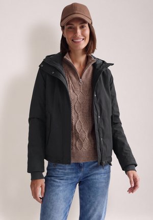 Femme souriante portant une casquette marron, une veste à capuche noire sur un pull en maille marron et un jean bleu, debout devant un fond clair uni.