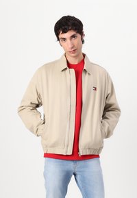 Beige zip-jakke med spiss krage, elastiske mansjetter og hem. Har liten brodert logo på venstre bryst. Bær over en rød genser.