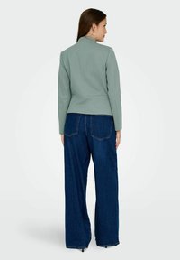 Femme vue de dos portant un blazer vert clair, un jean large foncé, des boucles d'oreilles anneaux et des talons noirs à bout pointu sur un fond clair uni.