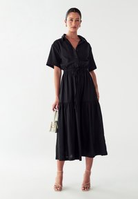 Abito lungo in cotone nero con maniche corte, apertura frontale con bottoni e vita cintrata. Presenta una gonna a strati e una texture morbida. La modella tiene una piccola borsa a mano.