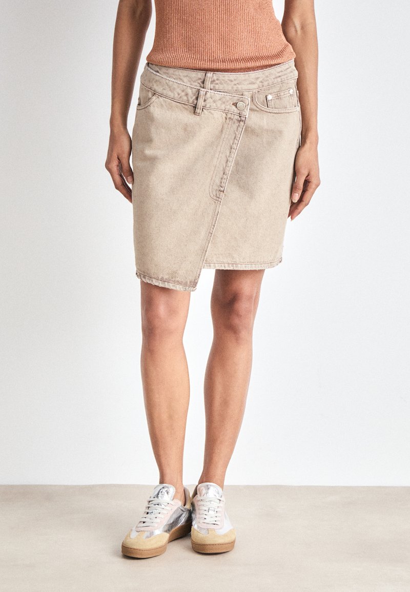 Gonna di jeans color beige chiaro con dettagli delle cuciture frontali, orlo leggermente curva e passanti per cintura. Indossata con sneaker grigie e beige.