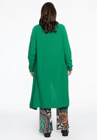 Groene lange cardigan van zachte stof, met lange mouwen en een ontspannen pasvorm, gecombineerd met meerkleurige, geprinte wijde broek en zwarte schoenen.