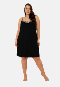 Kobieta w rozmiarze plus, stojąca boso, ubrana w czarną sukienkę bez rękawów z leoparda w wykończeniu dekoltu.