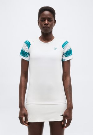 Persoană care poartă o rochie sportivă albă cu modele geometrice turcoaz pe mâneci scurte, stând pe un fundal simplu și deschis la culoare.