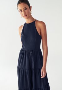 Mladá žena s tmavými vlasmi zviazanými vzadu, oblečená v bezrukávových modro-navy variabilných šatách a perlovými náušnicami, stojaca proti jednoduchému svetlému pozadiu.