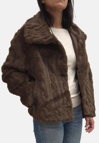 Giacca in pelliccia sintetica marrone con texture a maglia a trecce, colletto ampio, due grandi bottoni e tasche laterali con zip. Abbinata a una maglietta leggera e jeans.