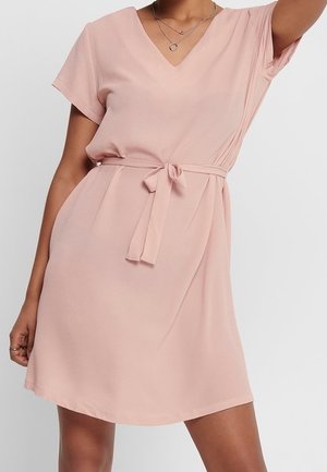 Kvinde iført en lys pink kjole med korte ærmer, V-hals og bånd i taljen, stående mod en ensfarvet lys baggrund.