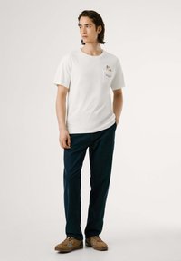 Witte korte mouwen t-shirt met een borstzak en grafische print, gecombineerd met marineblauwe broek en bruine schoenen met witte accenten.