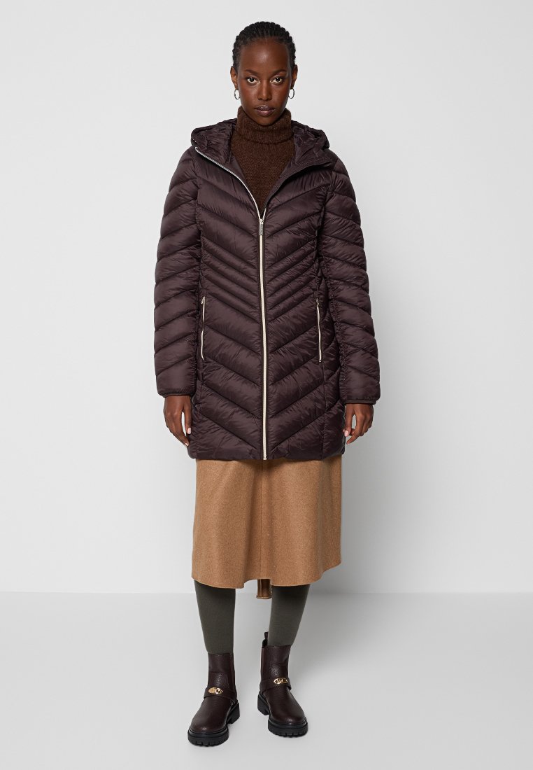 Ciemnobrązowa puchowa kurtka z kapturem, z przeszyciami w zygzak i zamek z przodu. Połączona z brązową spódnicą i ciemnymi butami.