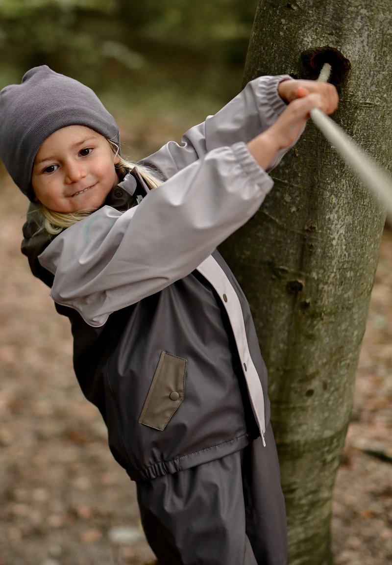 Enfant portant une veste d'extérieur grise et beige avec des poches, un bonnet gris, tirant une corde attachée à un arbre dans un environnement forestier.