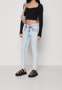 Žena v světle modrých skinny džínách, černém dlouhém rukávu zkráceném topu, černých sandálech na řemínky a béžové kabelce přes rameno na bílém pozadí.