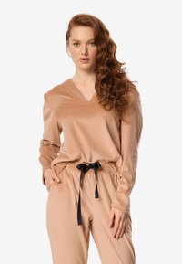 Satynowy, jasnobrązowy strój do domu z bluzką z dekoltem w serek oraz spodniami z elastycznym pasem i czarnym sznurkiem, o gładkiej fakturze i luźnym kroju.