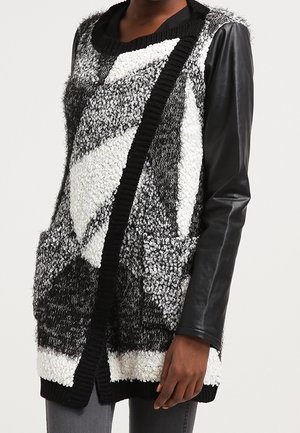 Zwart-witte gebreide cardigan met textuur en geometrisch patroon, zwarte geribbelde afwerking, voorzakken en lange zwarte leren mouwen, gedragen door een persoon.